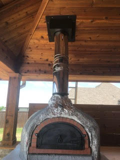 Outdoor kitchen stove rounded with concrete on outside and a black door under a beautiful grained wood ceiling